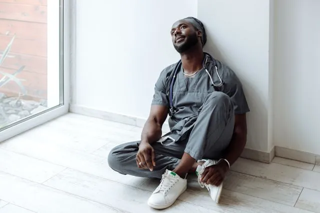 A doctor in scrubs sitting on the floor against a wall, taking a breath