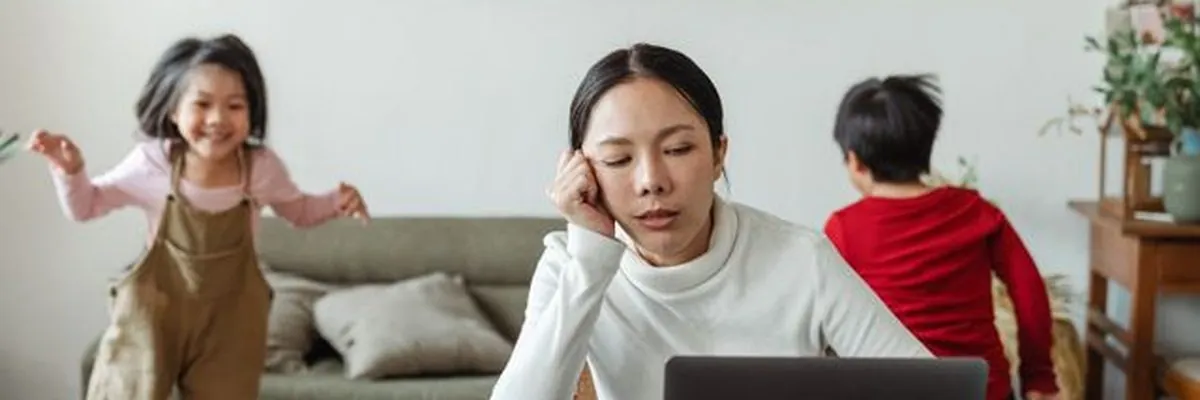 A tired mum working at her laptop while her children play behind her