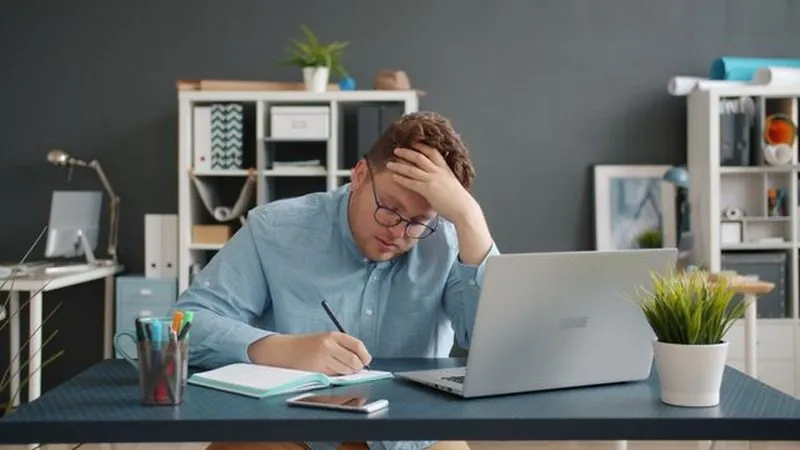 A tired teacher marking work at his desk beside a laptop