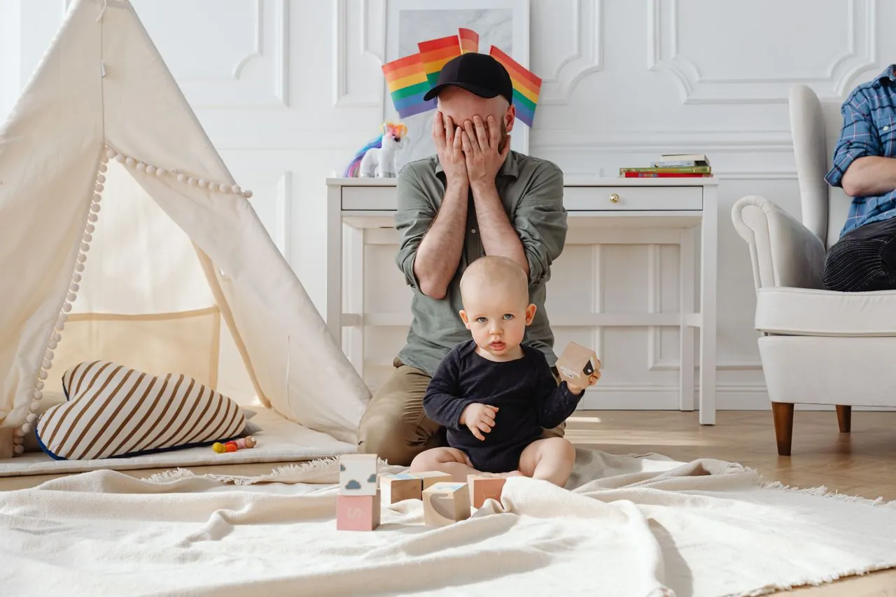 An overwhelmed dad covering his face while his baby sits playing with blocks